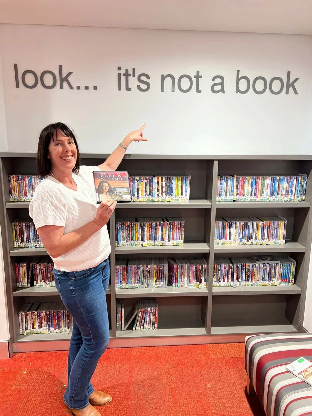 Fleur smiles and points to a sign above a shelf full of audiobooks. The sign reads: Look ... It's not a book. Fleur holds the audiobook of Broad River Station