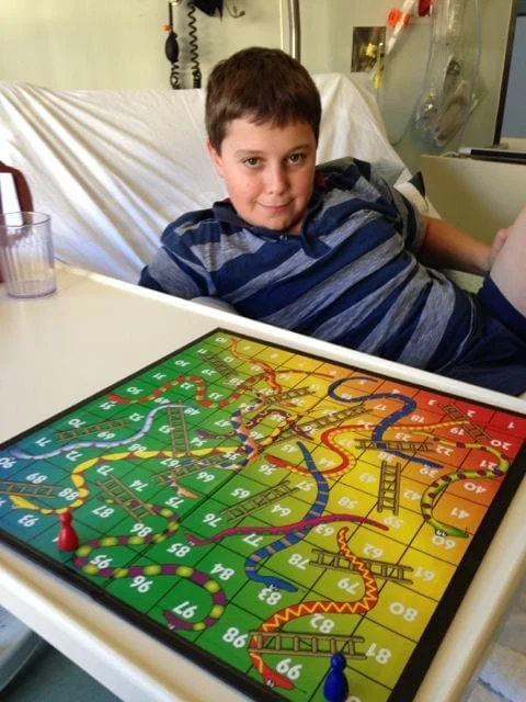 A young boy in a hospital bed, sitting up playing snakes and ladders