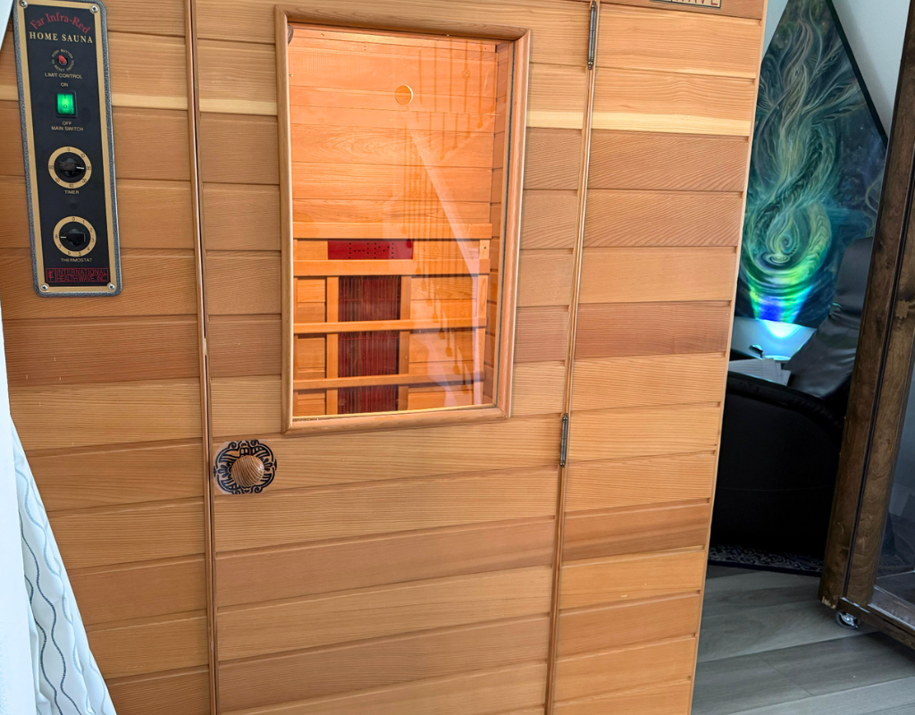 Close-up of a wooden infrared home sauna with a glass door and control panel. The control panel has switches and a timer, with a small vent on the wooden wall.