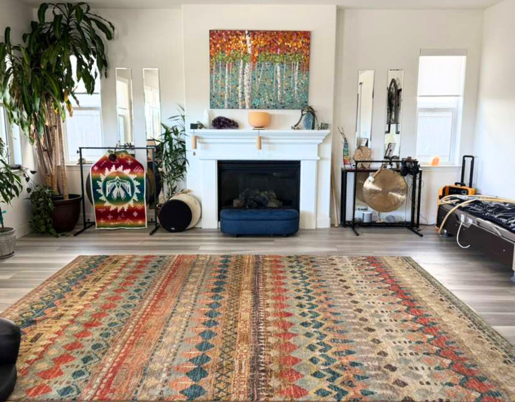 Living room with white walls, a colorful abstract painting above a white fireplace, green plants, and assorted decorative items. There is a large patterned rug on the floor, and windows on either side of the fireplace. Some furniture and objects, inc