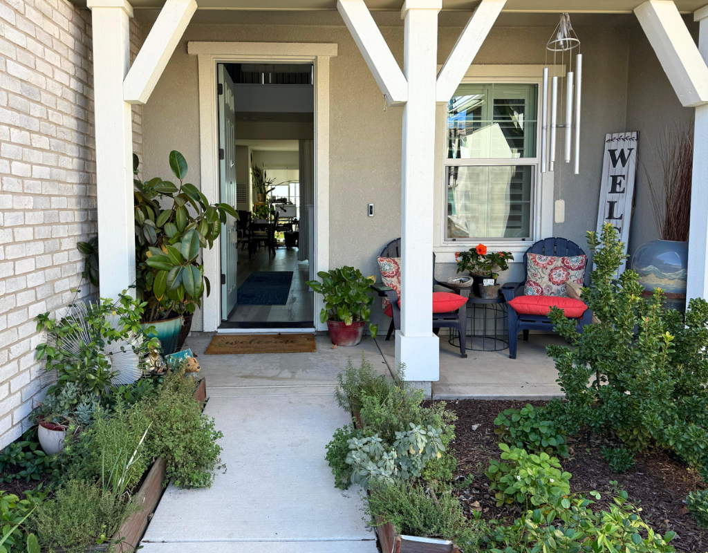 Covered front porch with two chairs with red cushions, potted plants, a wind chime, and a welcome sign.