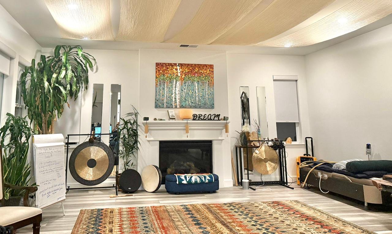 Living room with a white fireplace, decorative objects, and plants, musical gongs, a colorful rug, a window, and a bed or lounge chair on the right side.