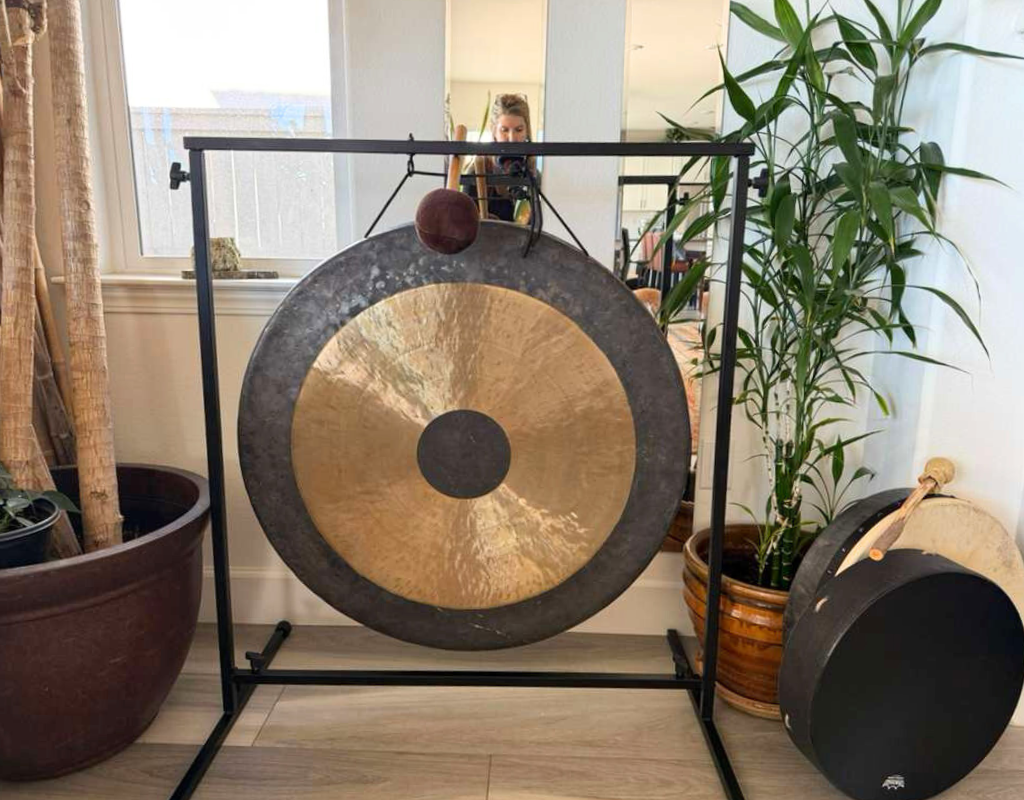 Giant gong with a metallic gold central circle, hung on a black metal stand indoors, with a woman taking a photo of it in the background, surrounded by potted plants.