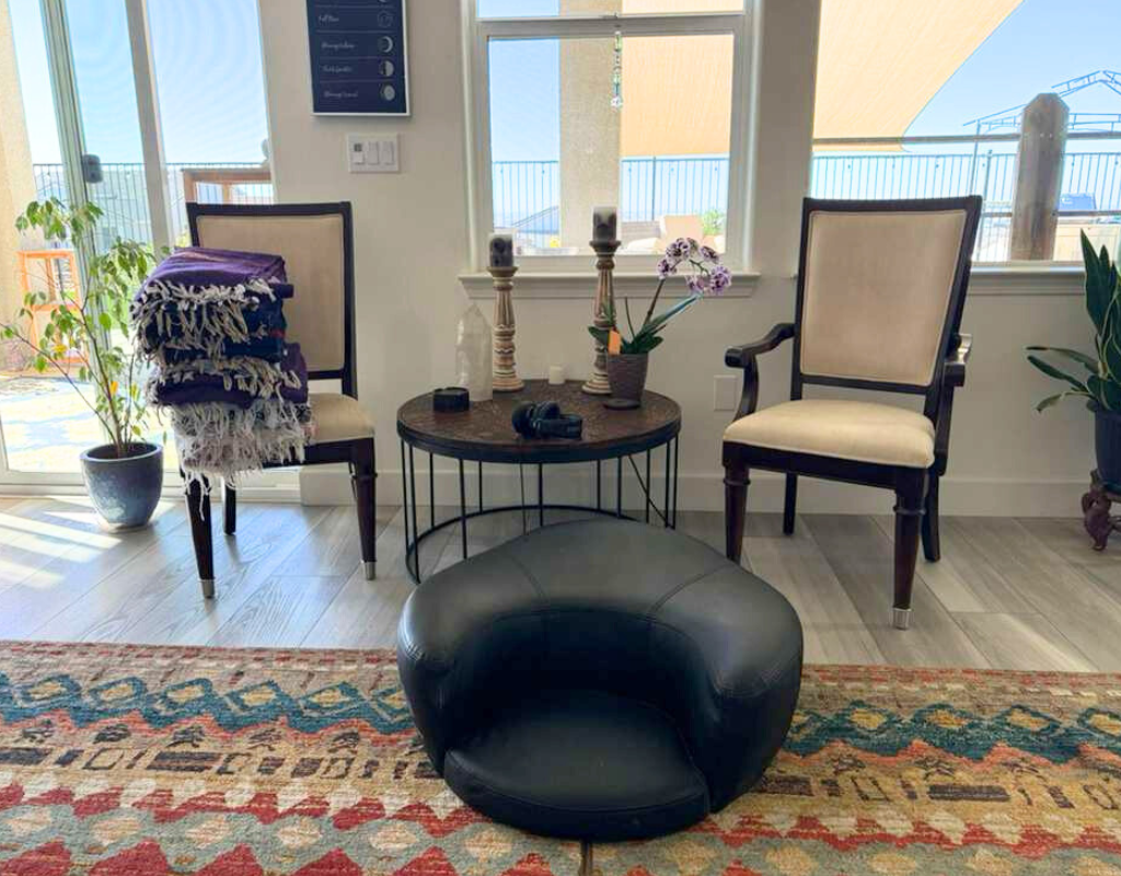 Living room with two beige armchairs, a small round wooden table with candles, a potted orchid, and a black leather ottoman. A stacked blanket is draped over one armchair, and there are potted plants near the sliding glass doors leading to a balcony.