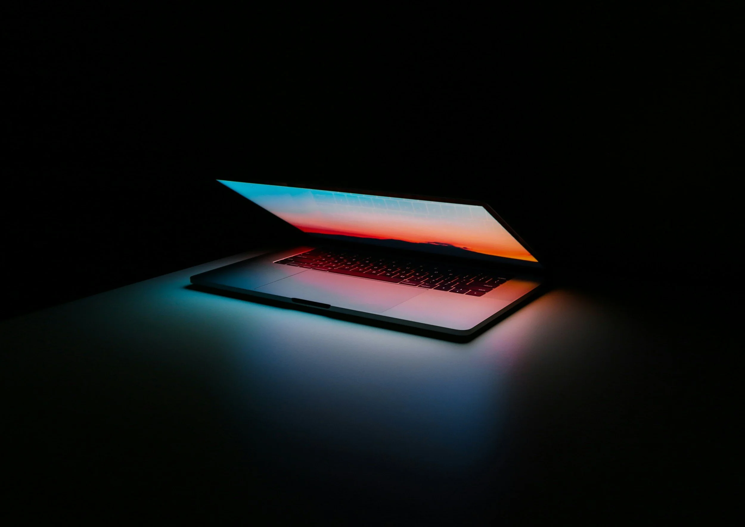 A laptop computer with a glowing screen displays a sunset landscape, in a dark room.