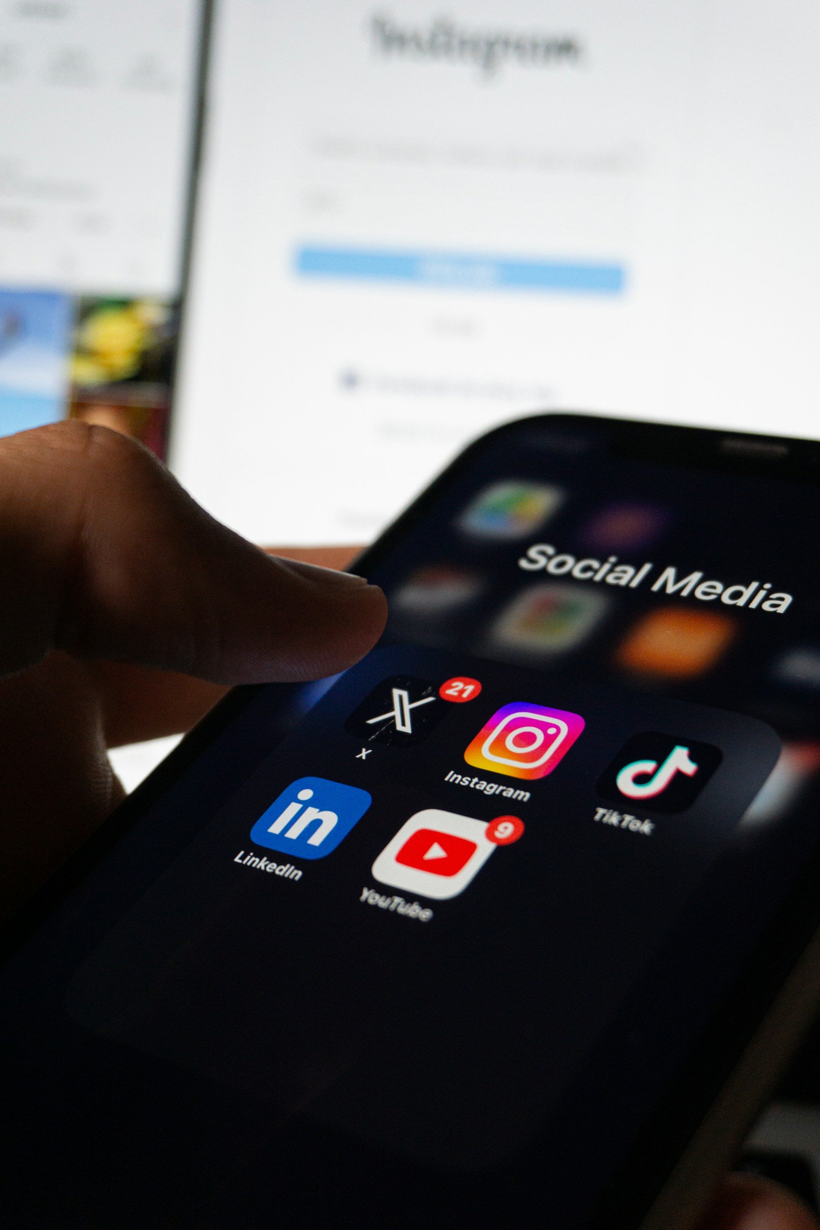 Close-up of a person's hand holding a smartphone with social media apps on the screen, including LinkedIn, YouTube, Instagram, TikTok, and an app with a black icon and red notification badge, with a blurred background showing a larger screen with the Instagram app open.