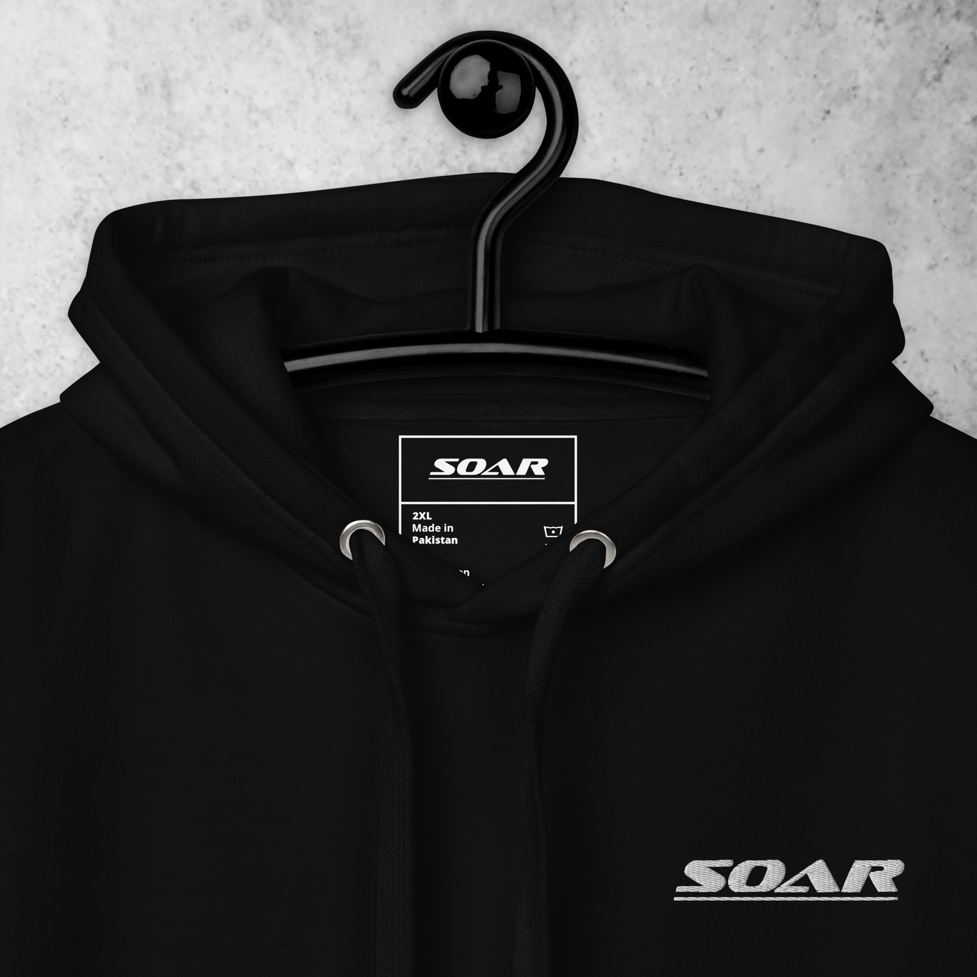Black hoodie on a hanger with a 'SOAR' logo on the chest, hood drawstrings, and a clothing label showing size 2XL, made in Pakistan, hanging against a gray background.
