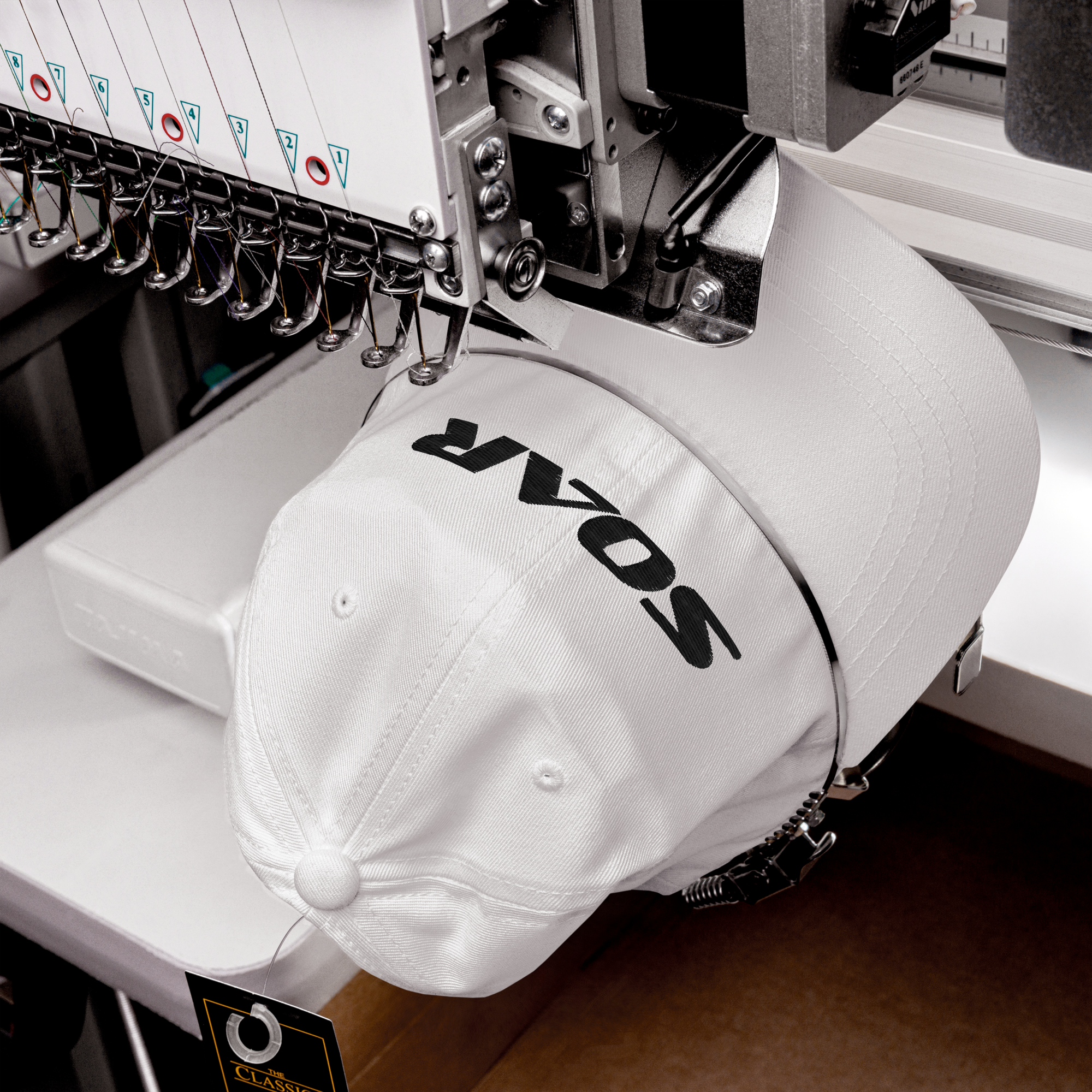 A white baseball cap with black lettering being embroidered on a commercial embroidery machine.