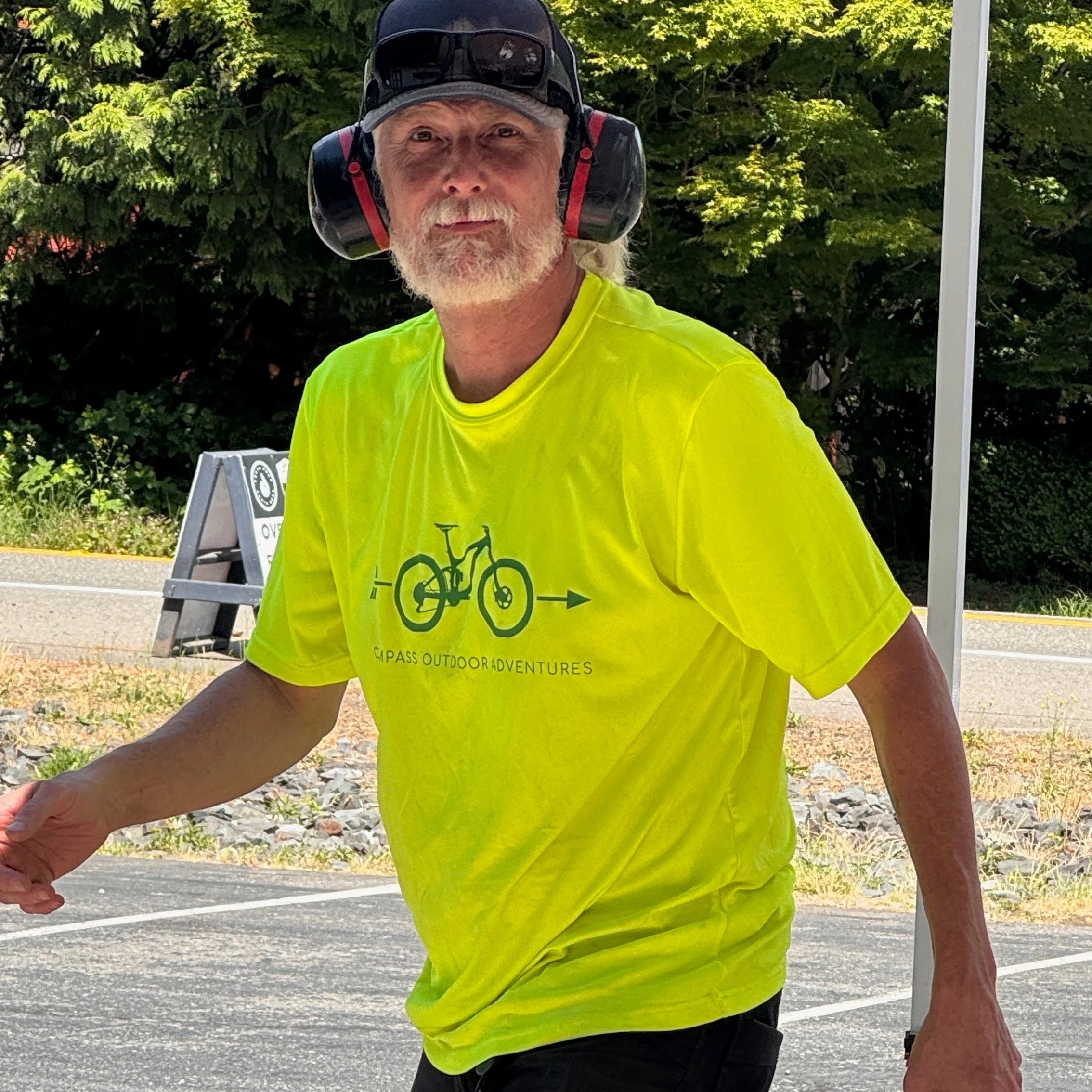A man wearing a bright yellow t-shirt, a black helmet with ear protection, and sunglasses, standing outdoors on a sunny day with green trees in the background.