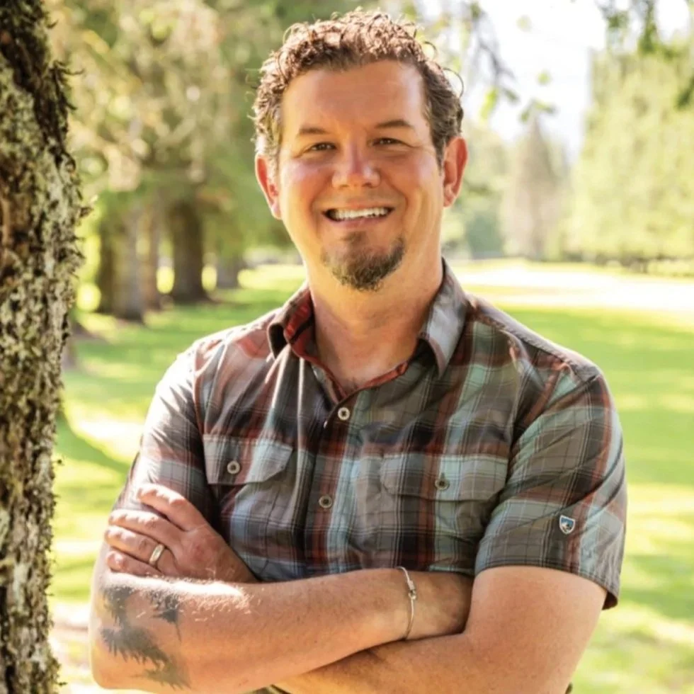A man with short curly hair and a goatee, smiling with arms crossed, standing outdoors near a tree in a park on a sunny day.