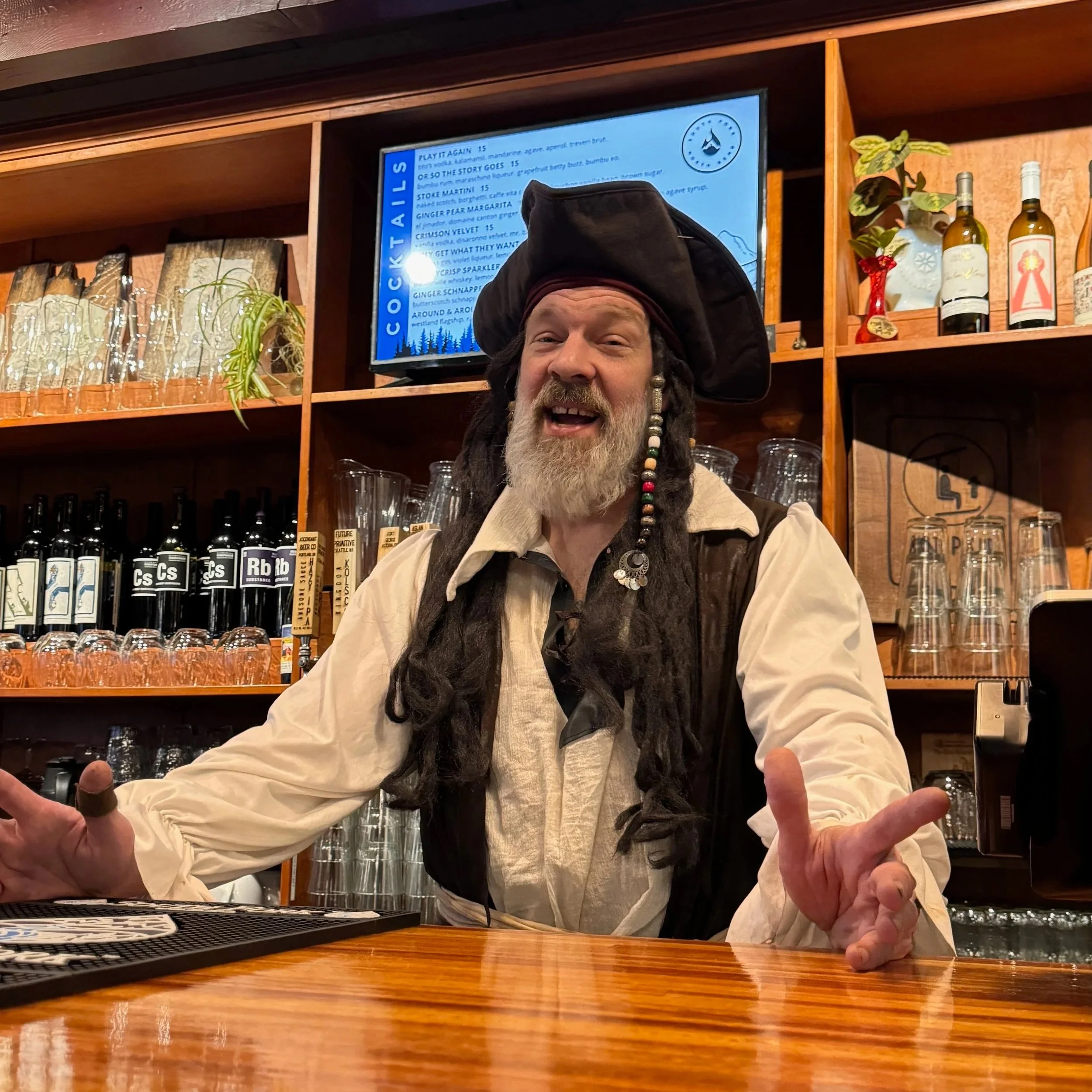 A man dressed as a pirate with long dreadlocks, a black pirate hat, and a white shirt with a brown vest, standing behind a wooden bar counter inside a bar or restaurant, smiling and gesturing with his hands.