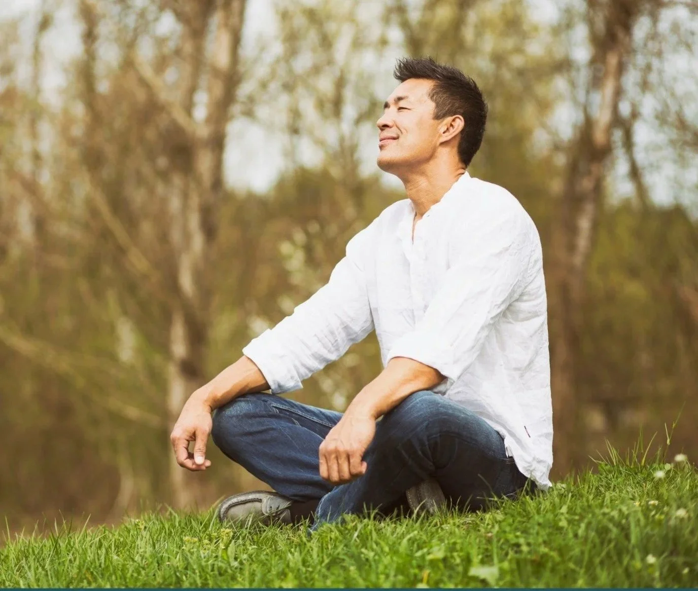 A man sitting cross-legged on grass with eyes closed, enjoying nature, surrounded by trees.