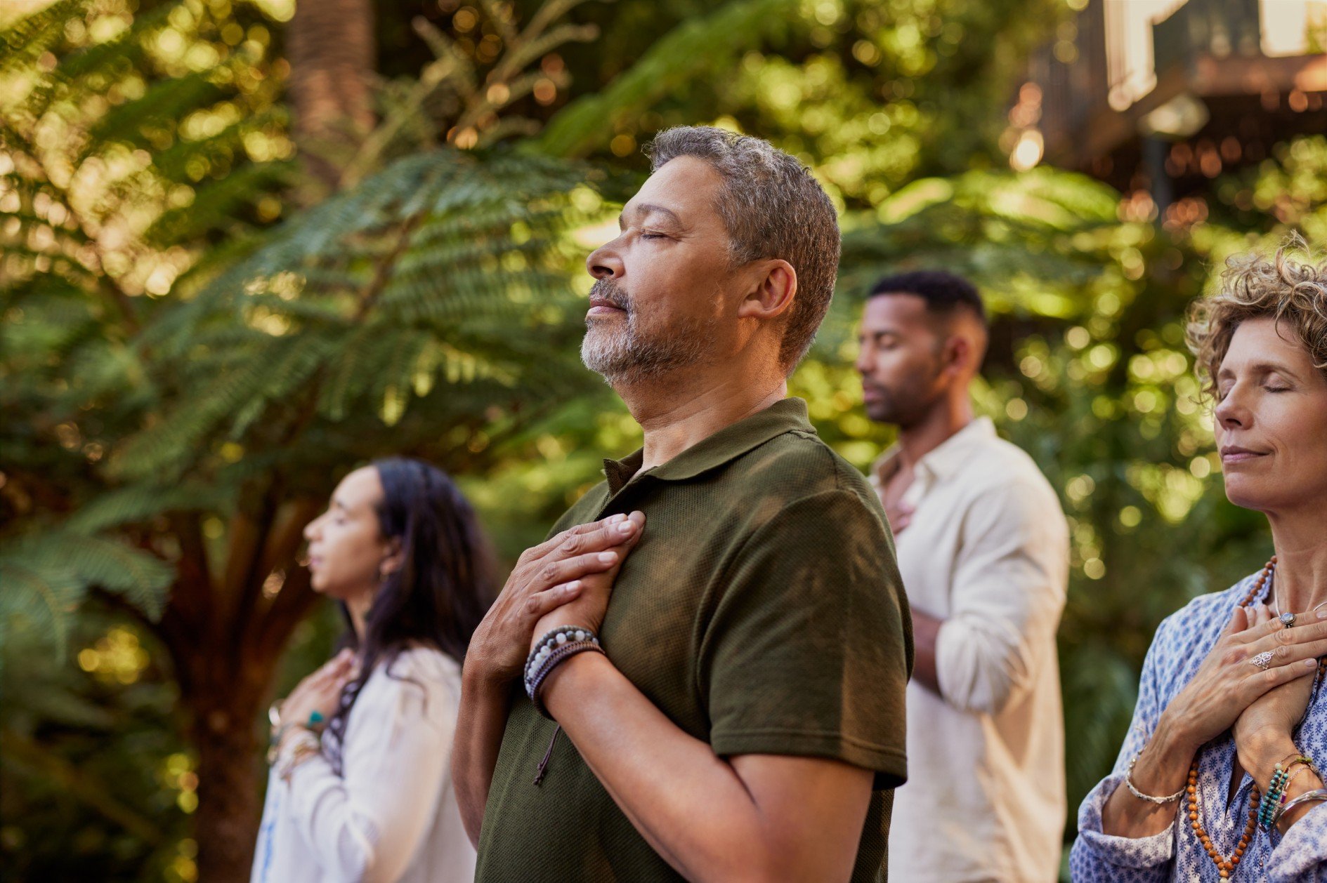 Four people standing outdoors with their eyes closed and hands over their hearts in a meditation or prayer pose, surrounded by green plants and trees.