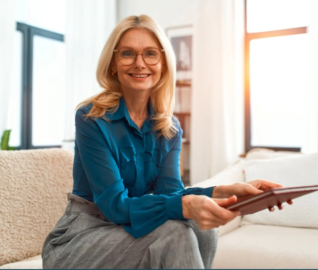 A smiling woman with blonde hair, glasses, wearing a blue blouse, sitting on a beige couch in a bright room with large windows, holding a tablet.