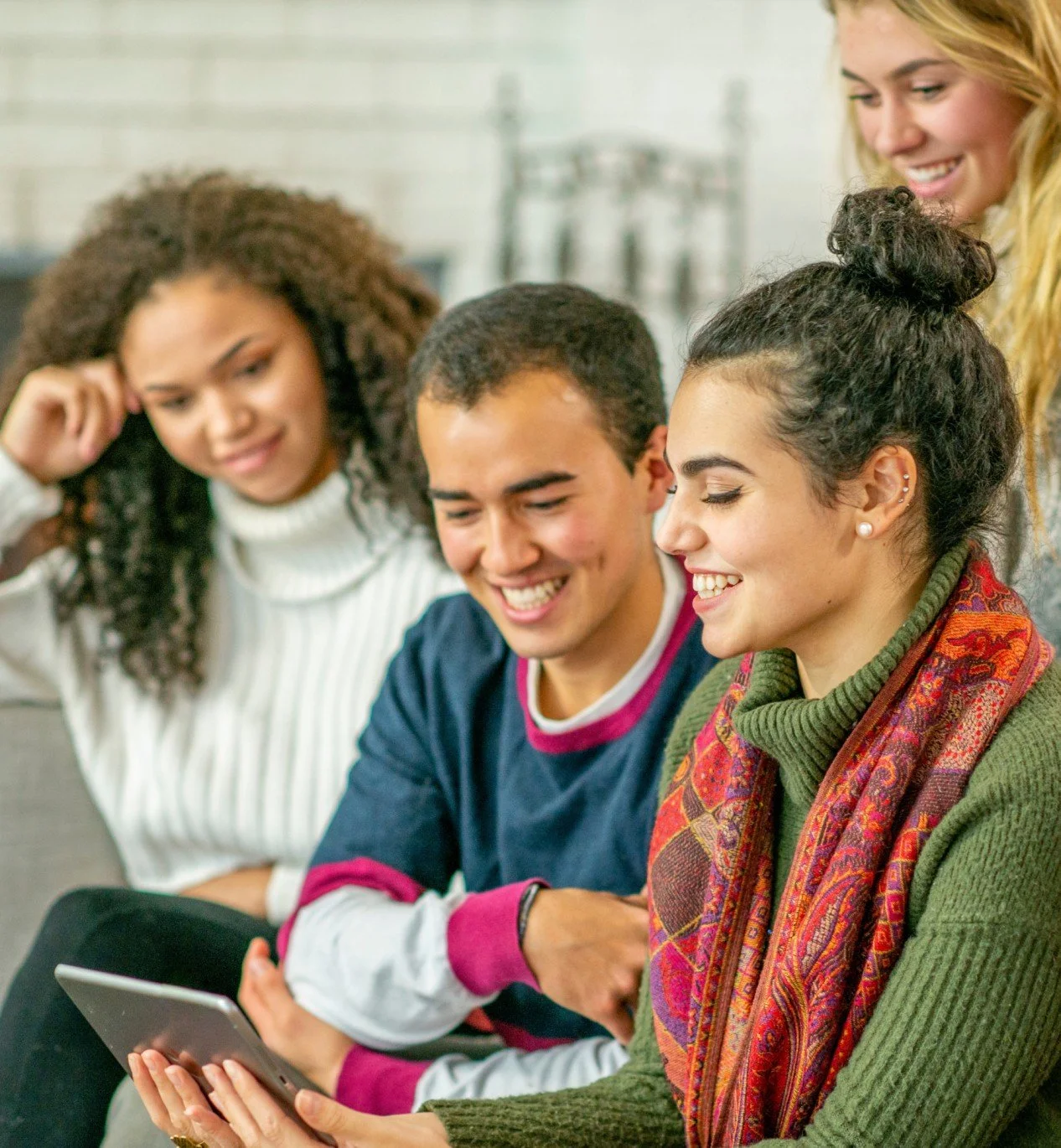 A group of diverse young people gathered together looking at a tablet, smiling and enjoying each other's company.