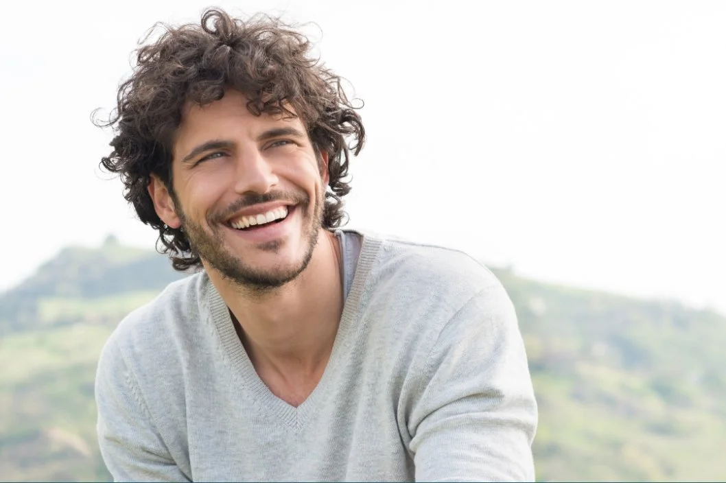 Smiling man with curly hair wearing a light gray sweater outdoors with a blurred hilly landscape in the background.