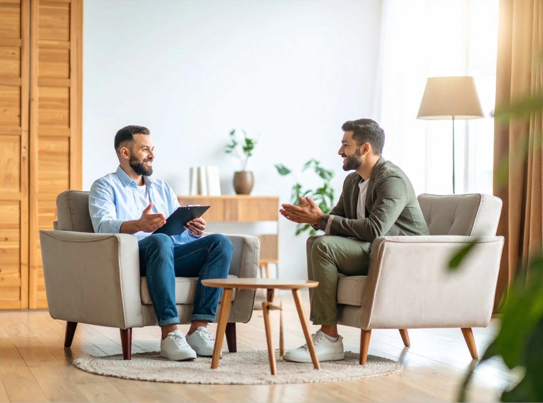 Two men sitting in armchairs and having a conversation in a well-lit living room with wooden furniture and green plants.