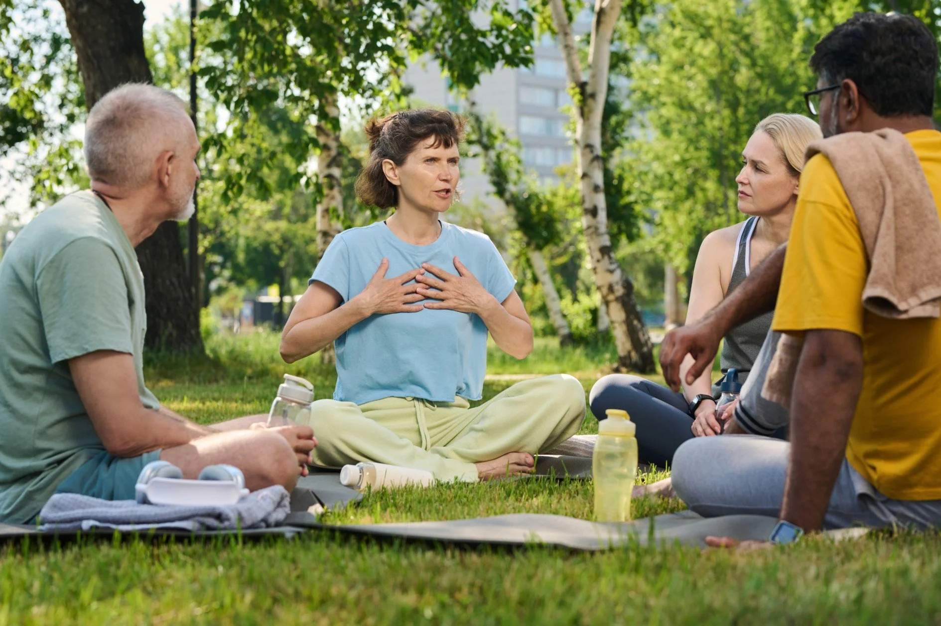 Group of people sitting on grass in a park, participating in a yoga or meditation session, with trees and buildings in the background.