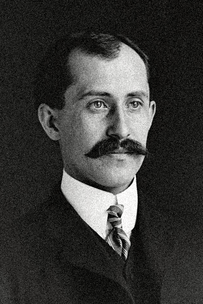 Black and white portrait of a man with a mustache, wearing a suit, tie, and white shirt.