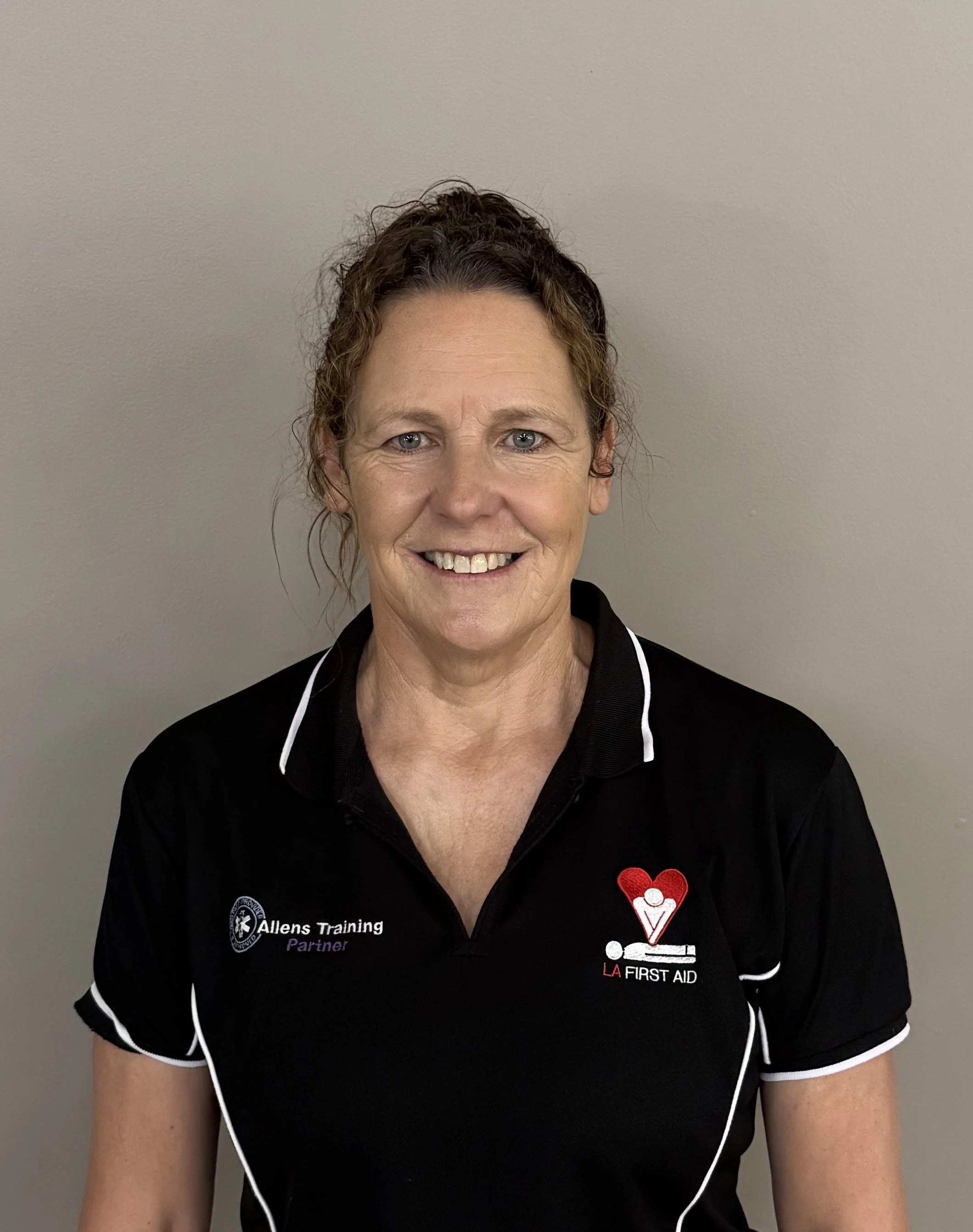 A woman smiling, wearing a black LA First Aid training shirt with white trim, standing against a plain gray wall.