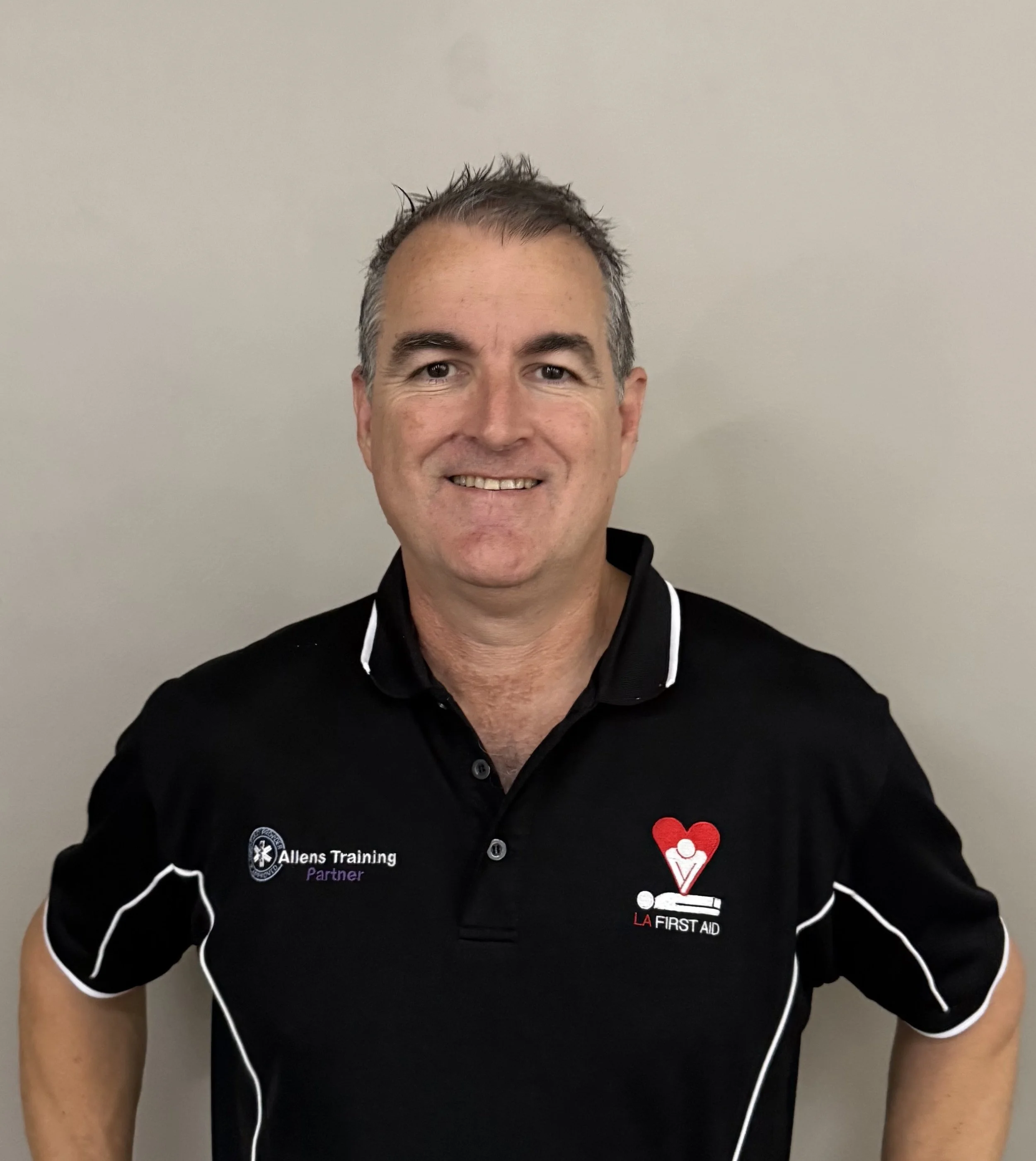 A man smiling at the camera, wearing a black polo shirt with logos for Allens Training and LA First Aid, standing against a plain light gray wall.