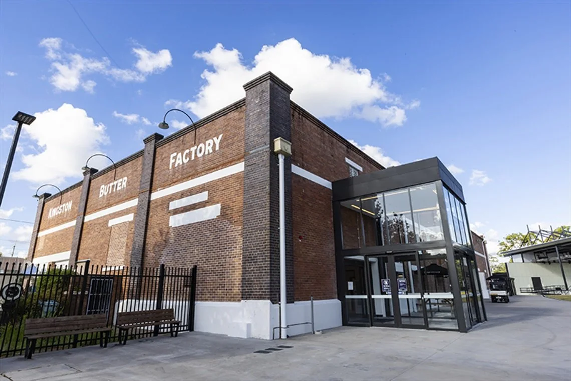 Exterior of a brick building labeled 'Kingston Butter Factory' with a glass entrance, black fence, benches, and a clear blue sky.