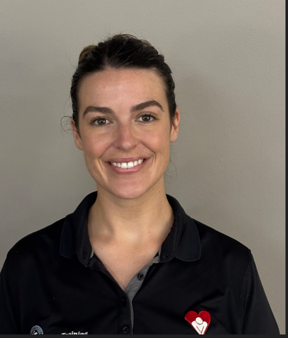 A woman with dark hair tied back, smiling, wearing a black polo shirt with a red heart logo on the chest, standing against a plain grey wall.
