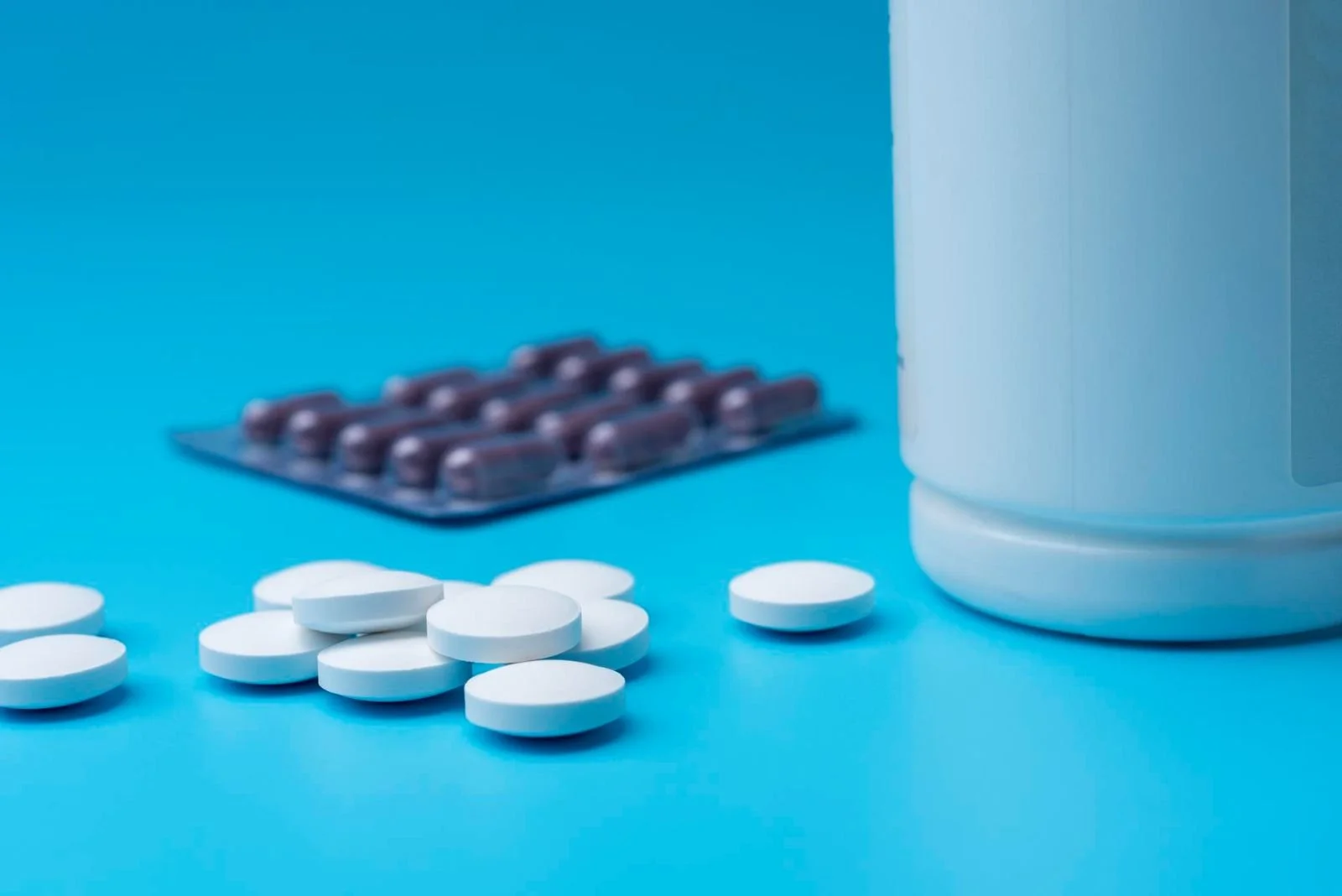 White round pills and a medicine bottle on a blue background with blister packs of purple capsules in the background.