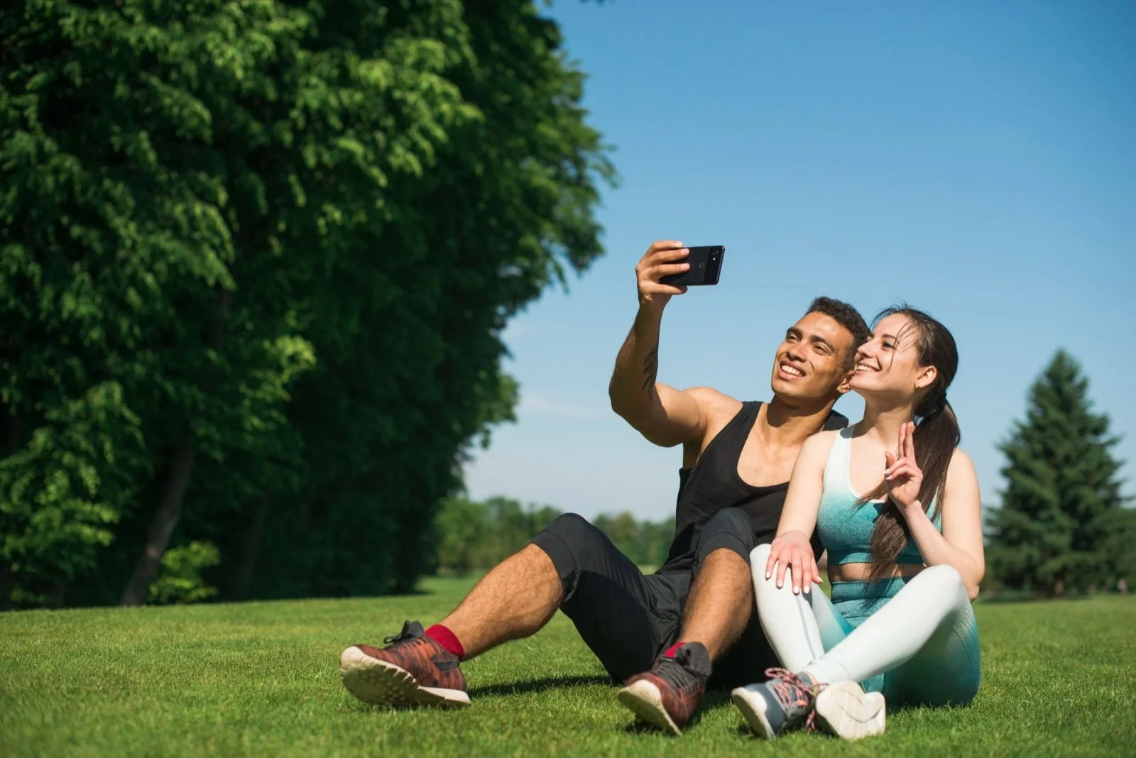 A man and woman sitting on grass in a park, taking a selfie together on a sunny day.