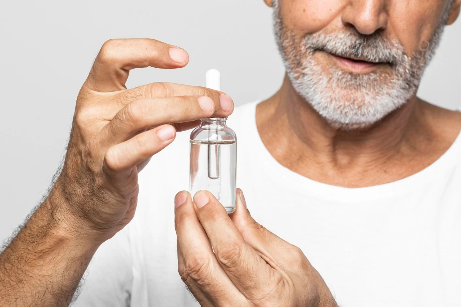 Close-up of a middle-aged man with a beard holding a small glass vial of clear liquid, with a white capsule pill on top, against a plain light background.