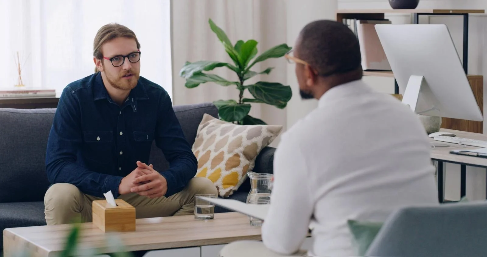 Two men having a conversation in a modern office or therapy room, with a plant and digital device in the background.