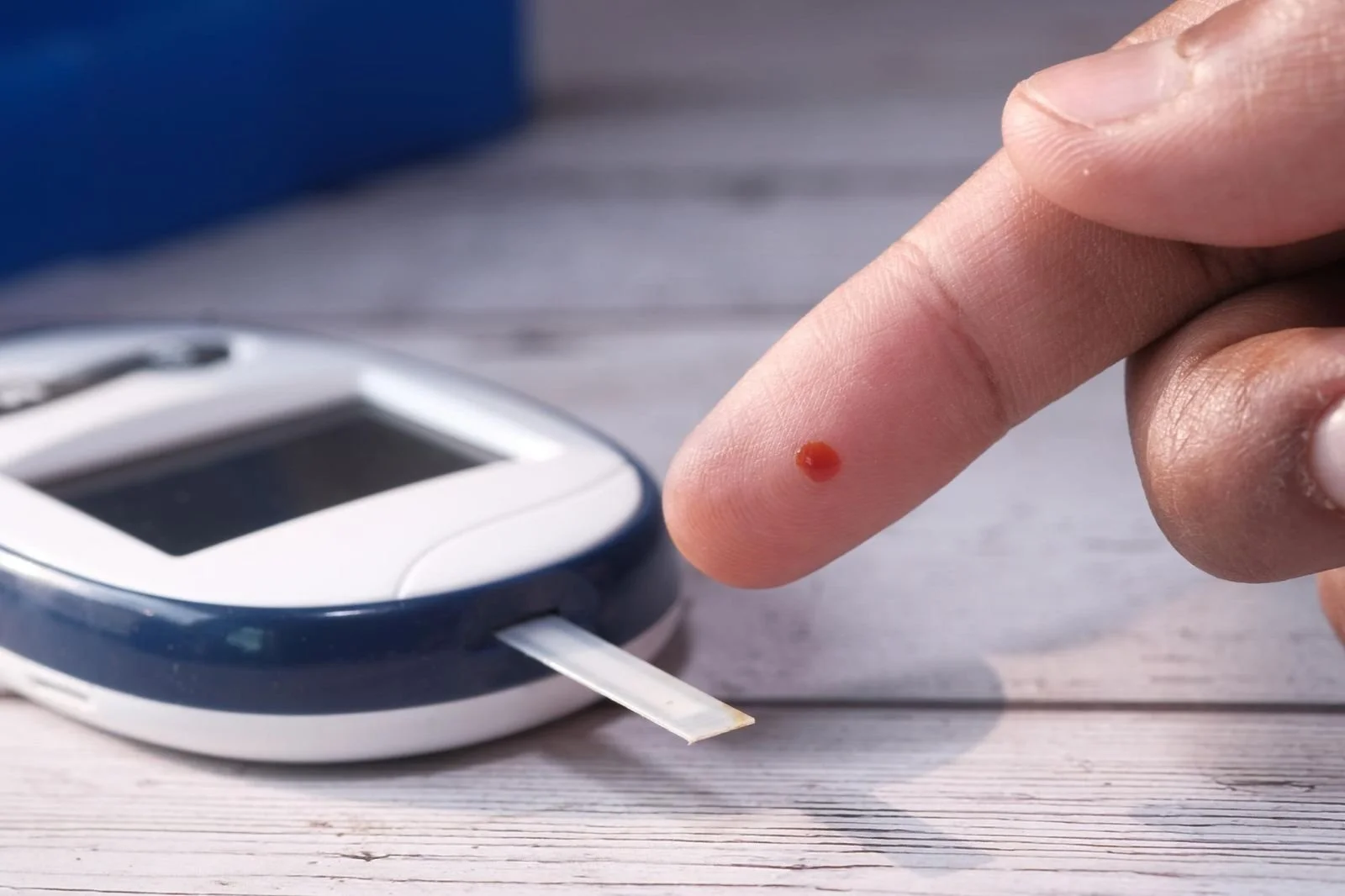 Close-up of a person's finger with a blood droplet near a glucometer on a wooden surface.