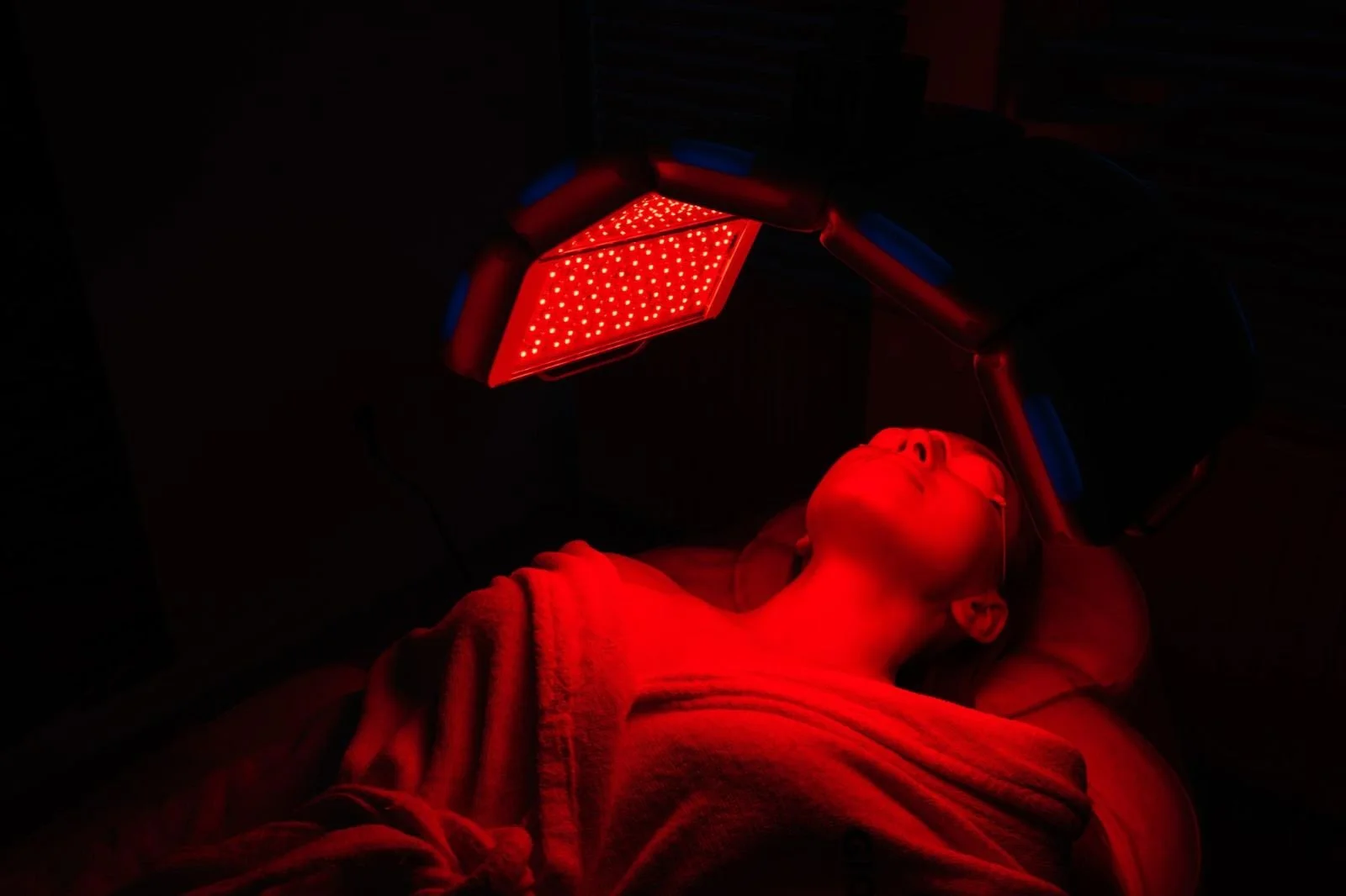 Person lying on a bed under red LED light therapy, with eyes closed and head tilted back.