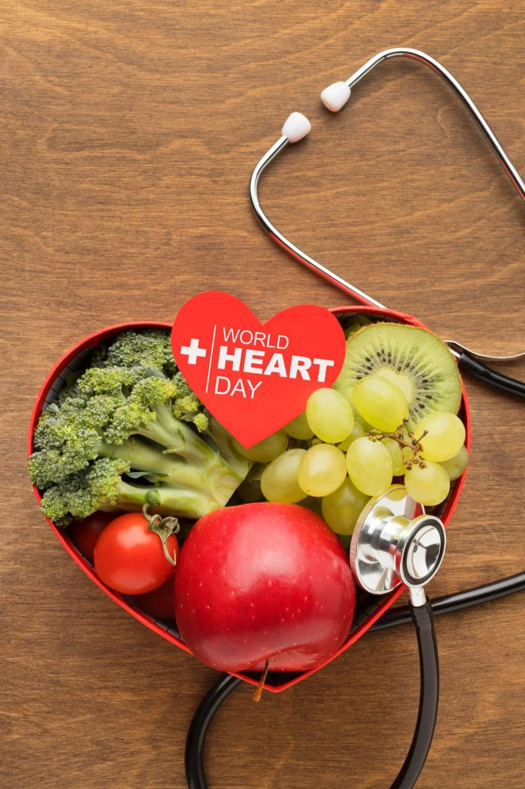 A heart-shaped box of green broccoli, a cluster of green grapes, a tomato, a red apple, and a slice of kiwi fruit, with a stethoscope and a 'World Heart Day' sign on a wooden surface.