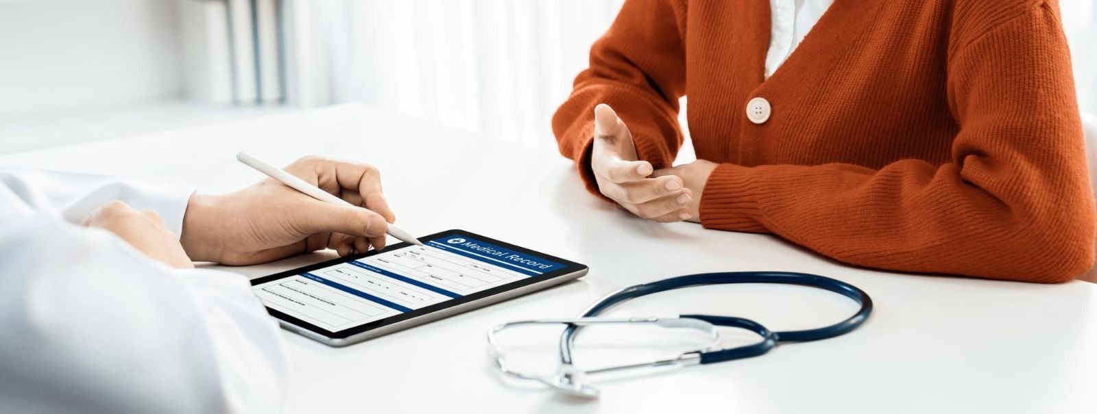 Doctor showing medical record on tablet to patient in a consultation room, with stethoscope on the table.