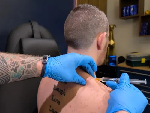 A person receiving a vaccination in their upper arm, administered by a healthcare professional wearing blue gloves, in a clinical setting.
