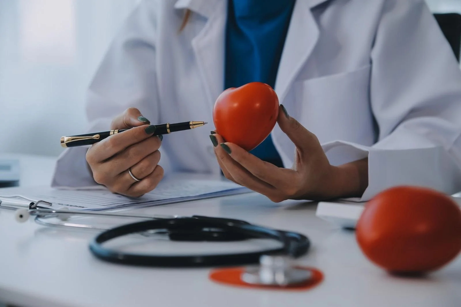 A healthcare professional wearing a white coat holding a tomato in one hand and gesturing with a pen in the other. A second tomato and a stethoscope are on the table.