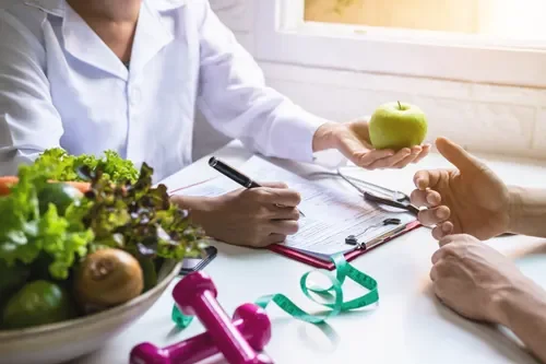 A healthcare professional consulting with a patient about nutrition, holding a green apple, with a bowl of fresh vegetables, pink dumbbells, and a measuring tape on the table.