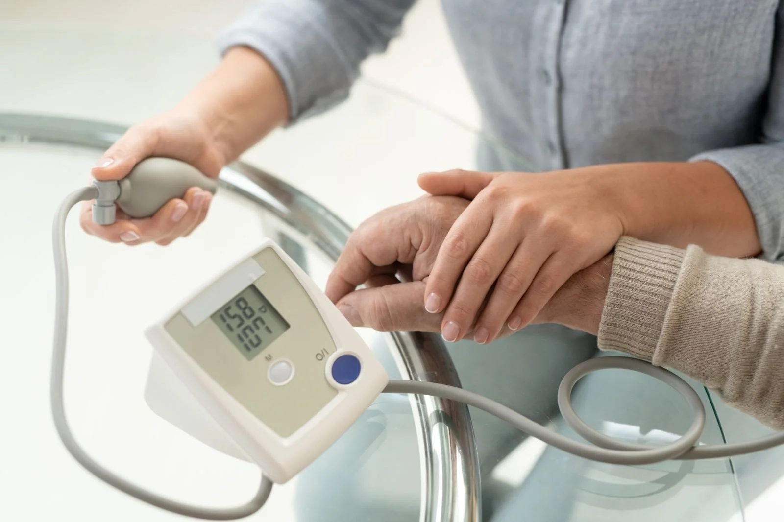 A person in gray sleeve measuring another person's blood pressure using a digital blood pressure monitor on a glass table.