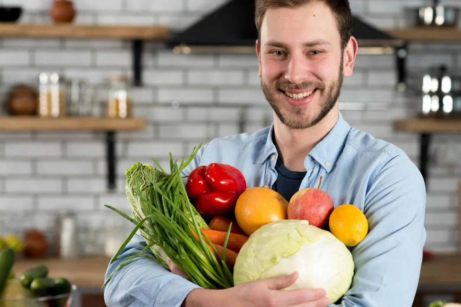 Smiling man holding a variety of fresh vegetables including lettuce, red bell pepper, orange, apple, lemon, cabbage, and carrots in a modern kitchen.