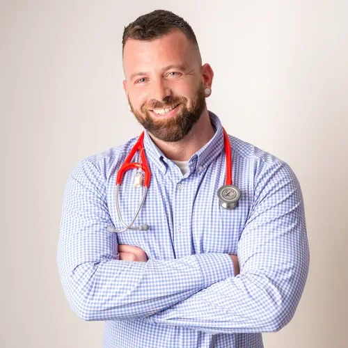 Portrait of a smiling male healthcare professional with a beard, wearing a light blue checkered shirt and a red stethoscope around his neck, standing with arms crossed against a plain background.
