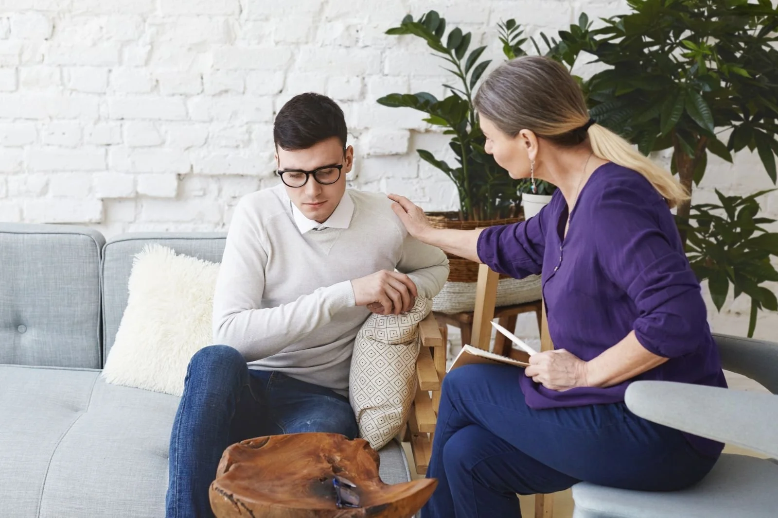 A young man with glasses sits on a sofa, looking distressed, with a woman in purple talking to him and touching his shoulder, in a bright room with a white brick wall and a large plant in the background.