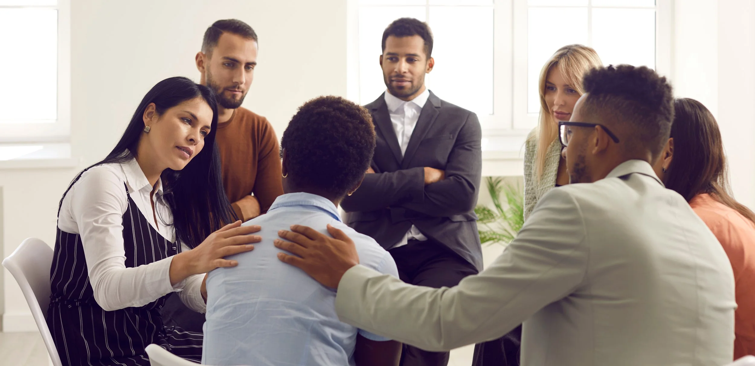 A diverse group of people engaged in a serious discussion in a bright, modern office space, with some members comforting or supporting one individual.