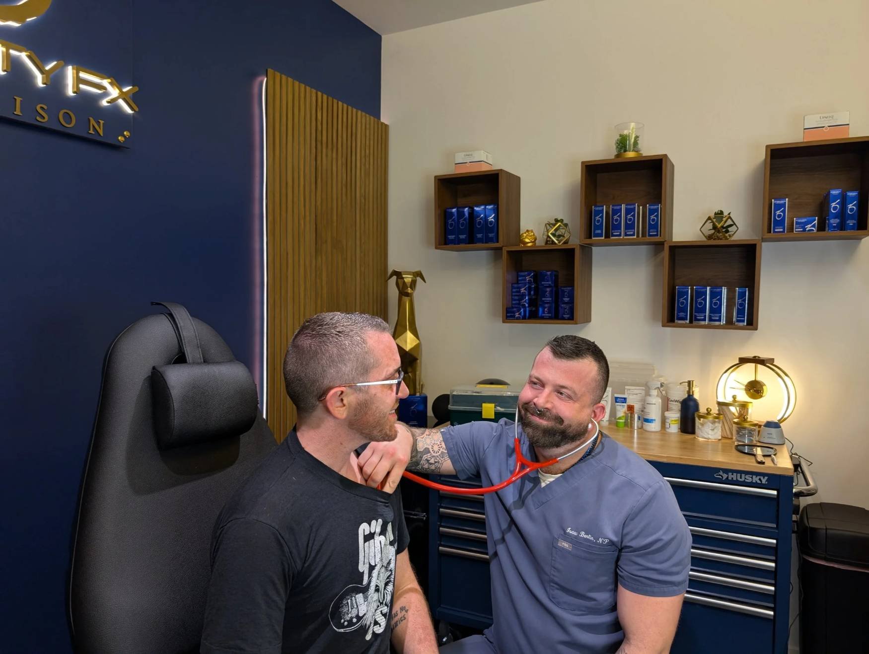 A healthcare professional examines a patient with a stethoscope in a medical office, with shelving displaying blue boxes and decorative items in the background.