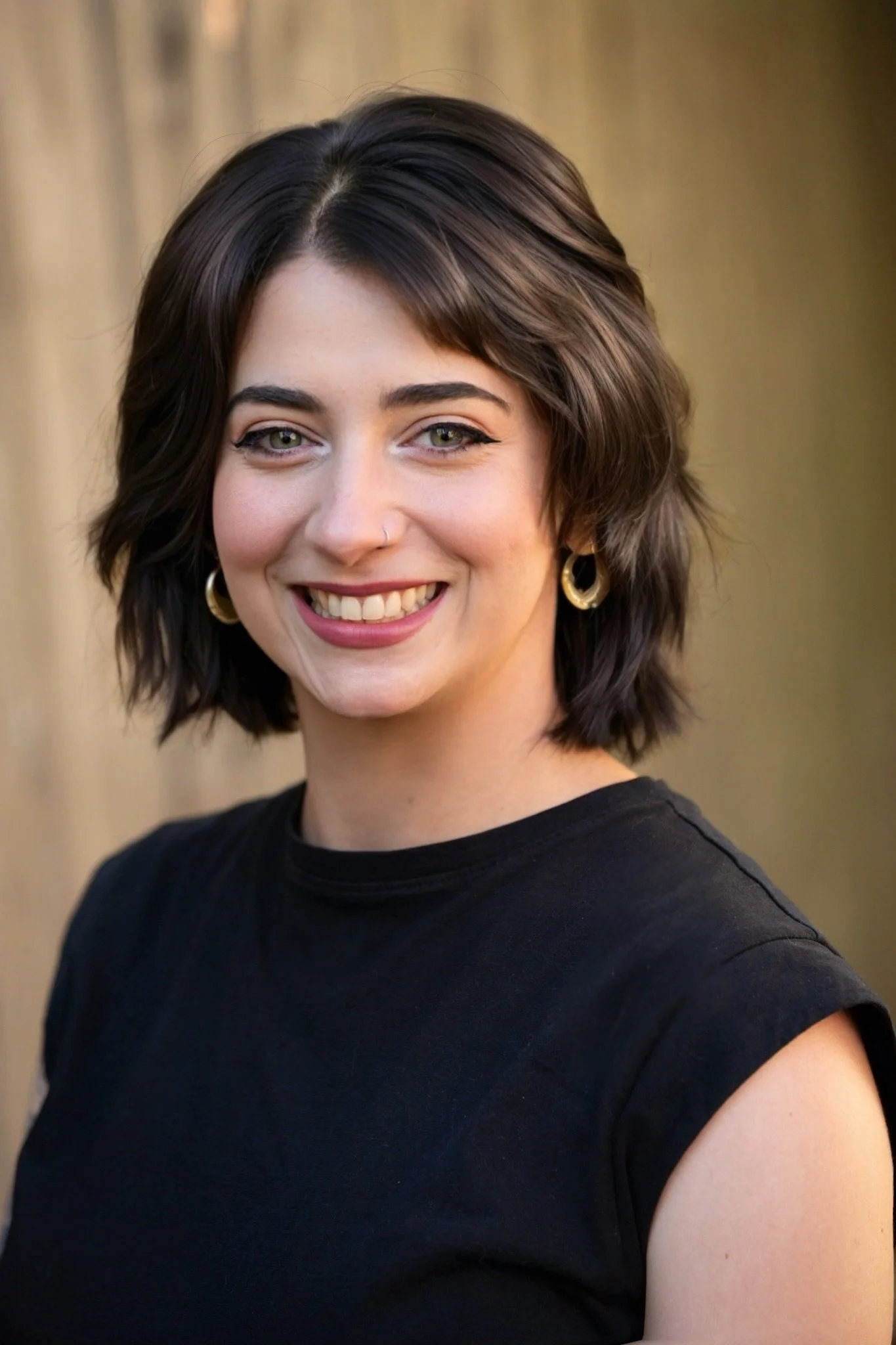 A woman with short dark brown hair, green eyes, and earrings, smiling at the camera, wearing a black top, outdoors with a blurred background.