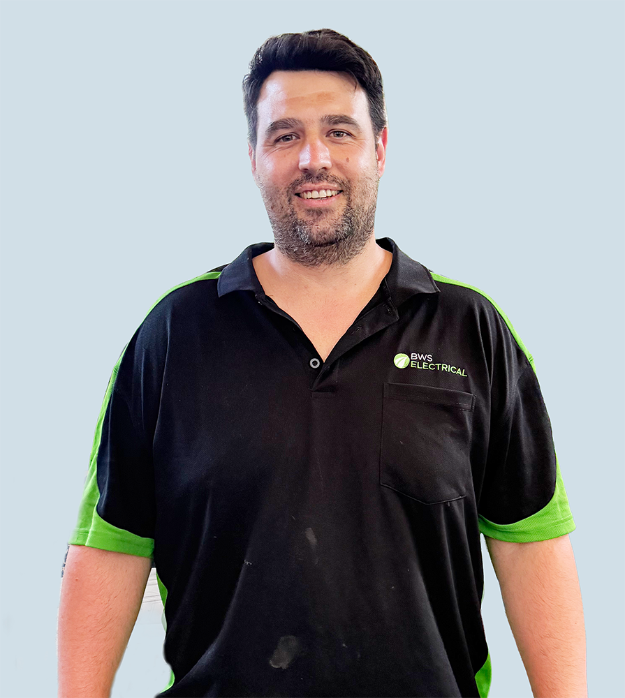 A man with dark hair and facial hair smiling, wearing a black and green company uniform with the logo 'BWS Electrical' on the shirt, standing against a light blue background.