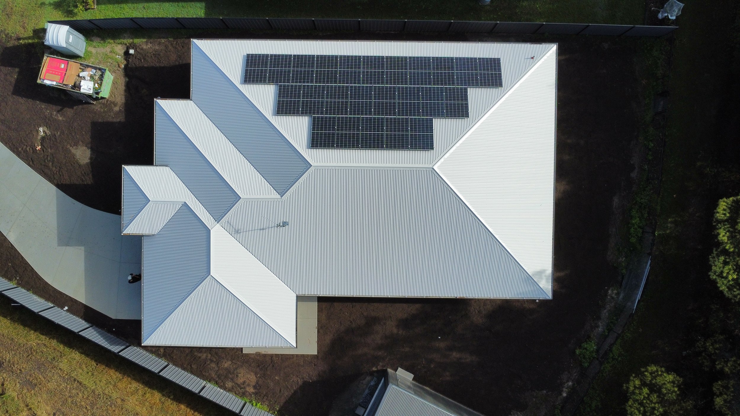 Aerial view of a house with a metal roof and solar panels, surrounded by a black fence, with a small white shed, green trees, and a driveway.