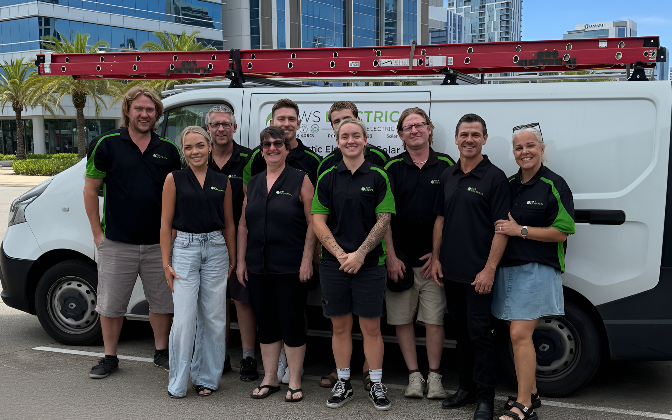 Group of nine people standing in front of a work van with a ladder on top, posing for a photo outdoors.