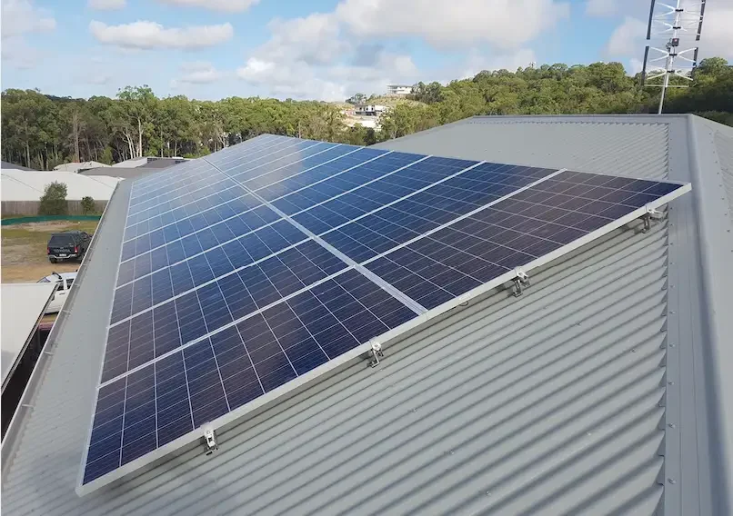 Solar panels installed on a slanted metal roof in a residential area with trees and a blue sky in the background.