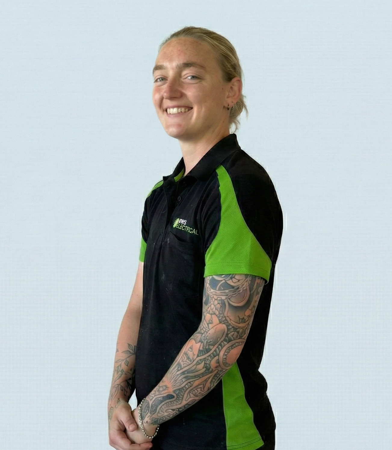 A smiling woman with blonde hair tied back, wearing a black and green polo shirt, standing against a plain light blue background, with tattoos on her forearms.